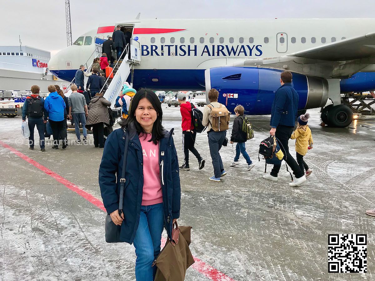 British Airways plane on the ground, woman with hand luggage standing in front of it. People boarding the plane.